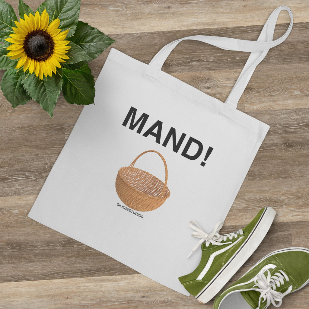 Witte MAND tote bag met opdruk van een mand en zwarte tekst liggend op houten vloer naast groene sneakers en zonnebloem