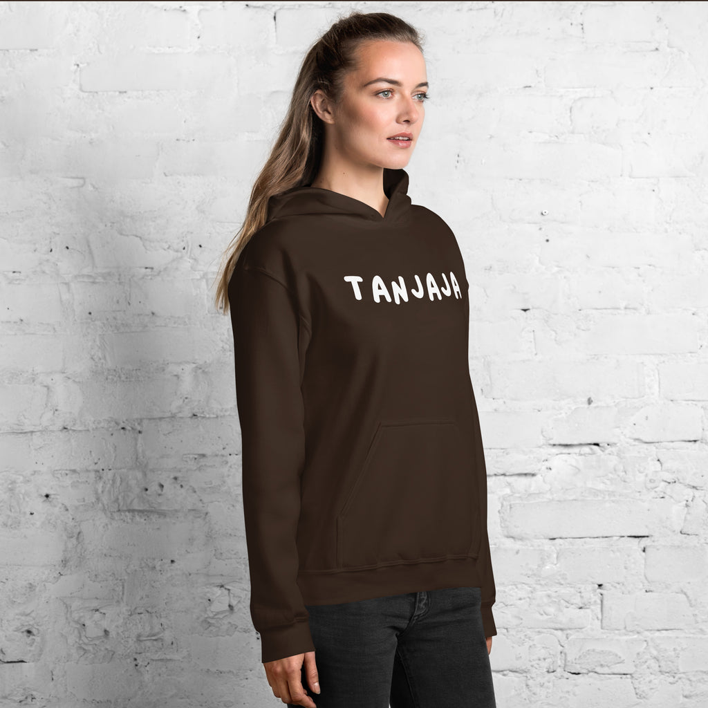 Vrouw draagt een bruine Tanjaja hoodie unisex met witte opdruk tegen een witte bakstenen muur